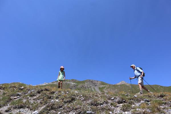 An adult and a child are seen hiking up a rocky, grassy mountain slope under a vast, clear blue sky